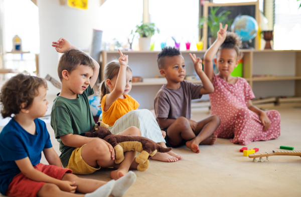 A group of small nursery school children sitting on floor indoors in classroom, raising hands.