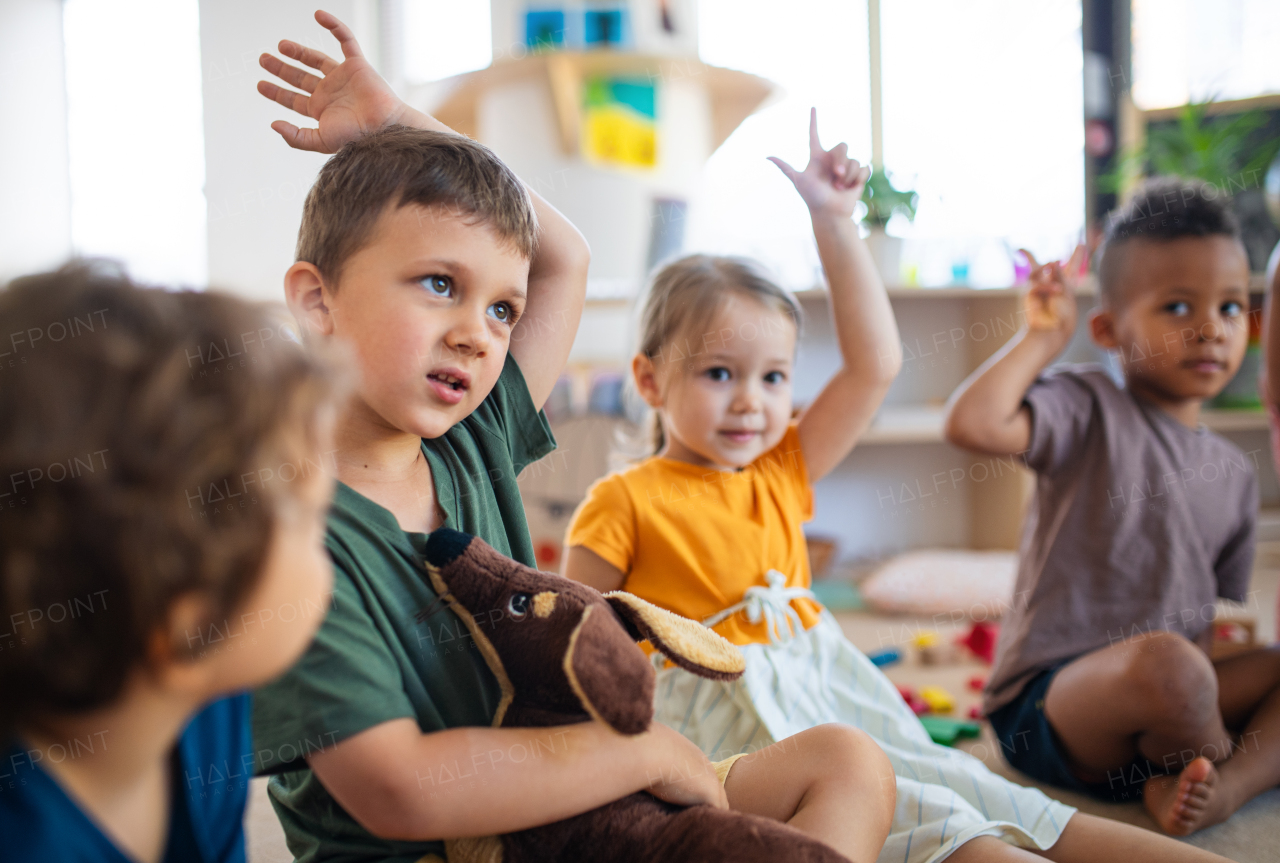 A group of small nursery school children sitting on floor indoors in classroom, raising hands.