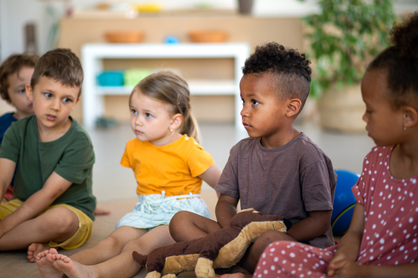 A group of small nursery school children sitting on floor indoors in classroom, listening to teacher.