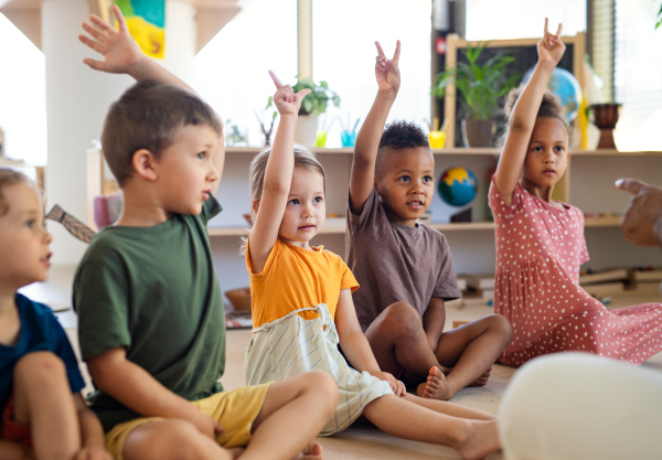 A group of small nursery school children sitting on floor indoors in classroom, raising hands.