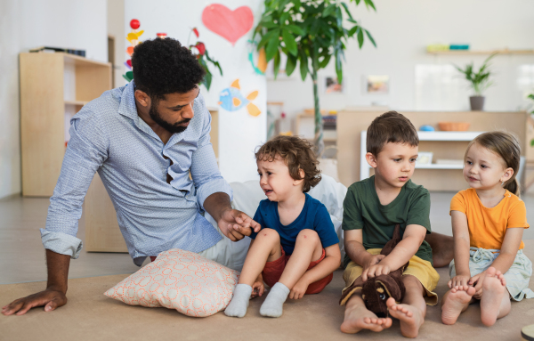 A man teacher talking to small crying boy indoors in classroom, comforting him.