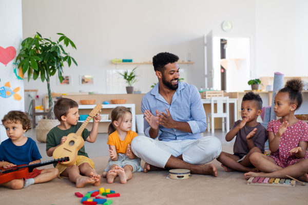 A group of small nursery school children with man teacher sitting on floor indoors in classroom, playing musical instruments.