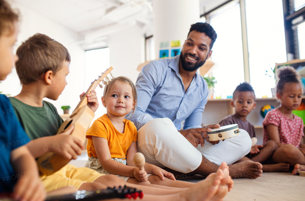 Group of small nursery school children with man teacher sitting on floor indoors in classroom, playing musical instruments.