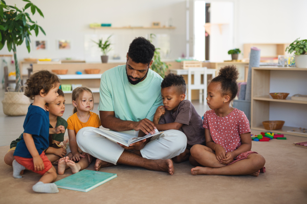 A group of small nursery school children with man teacher sitting on floor indoors in classroom, reading fairy tale book.