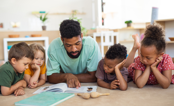 A group of small nursery school children with man teacher on floor indoors in classroom, reading book.