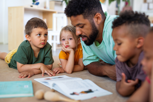 A group of small nursery school children with man teacher on floor indoors in classroom, reading book.