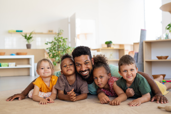 A group of small nursery school children with man teacher on floor indoors in classroom, looking at camera.