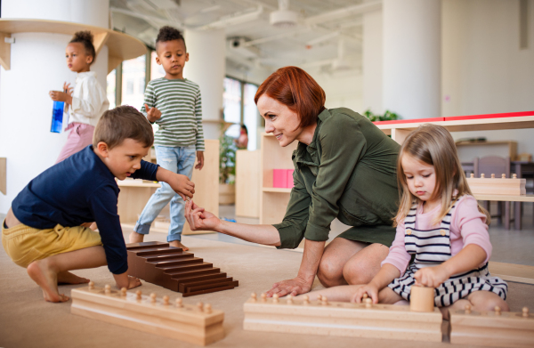 Group of small nursery school children with teacher sitting on floor indoors in classroom, montessori learning concept.