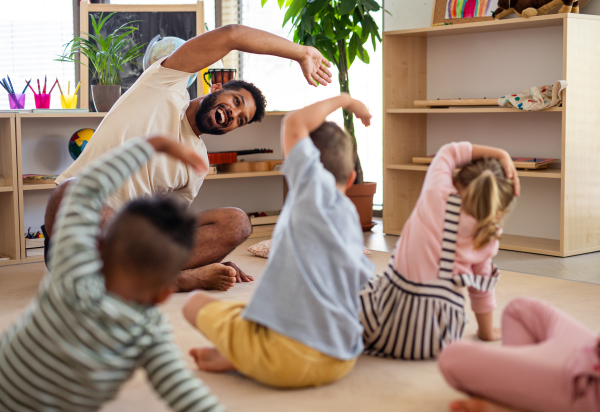 A group of small nursery school children with man teacher sitting on floor indoors in classroom, doing exercise.