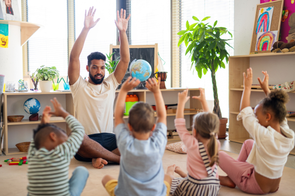 A group of small nursery school children with man teacher sitting on floor indoors in classroom, doing exercise.