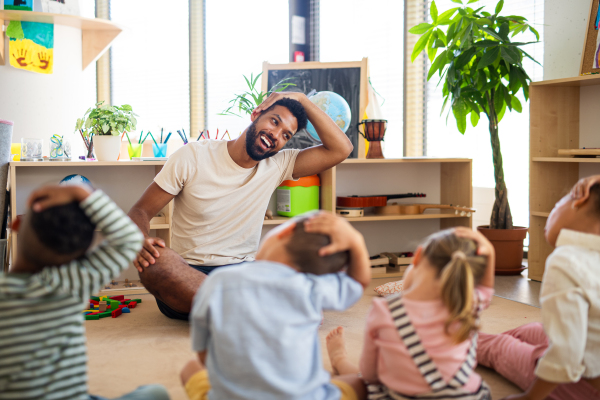 A group of small nursery school children with man teacher sitting on floor indoors in classroom, doing exercise.