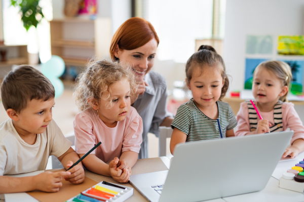 A group of small nursery school children with teacher on floor indoors in classroom, using laptop.