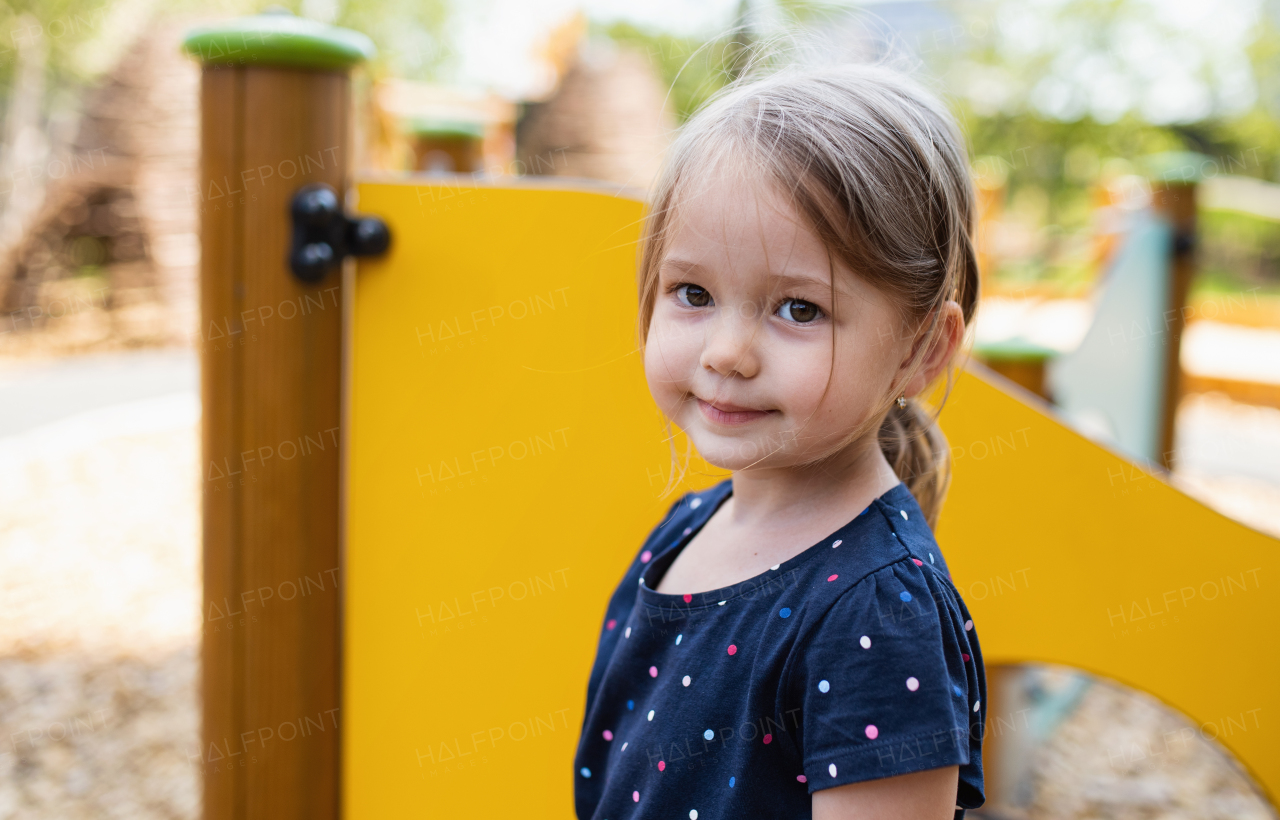 Small cute nursery school girl standing outdoors on playground, looking at camera.