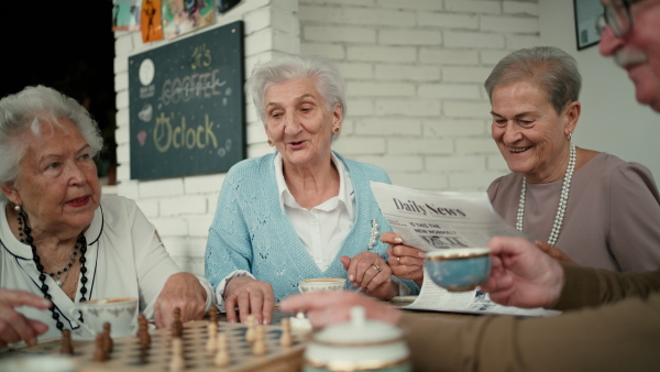 Happy senior friends sitting indoors in a cafe drinking coffee or tea, playing chess laughing and having nice time together.