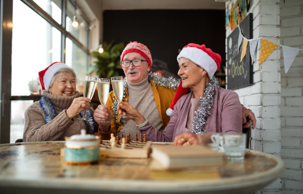 Happy senior friends sitting indoors in a cafe clinking champagne glasses and celebrating Christmas.