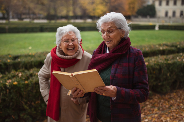 Happy senior women friends on a walk outdoors in town park in autumn, reading book and talking.