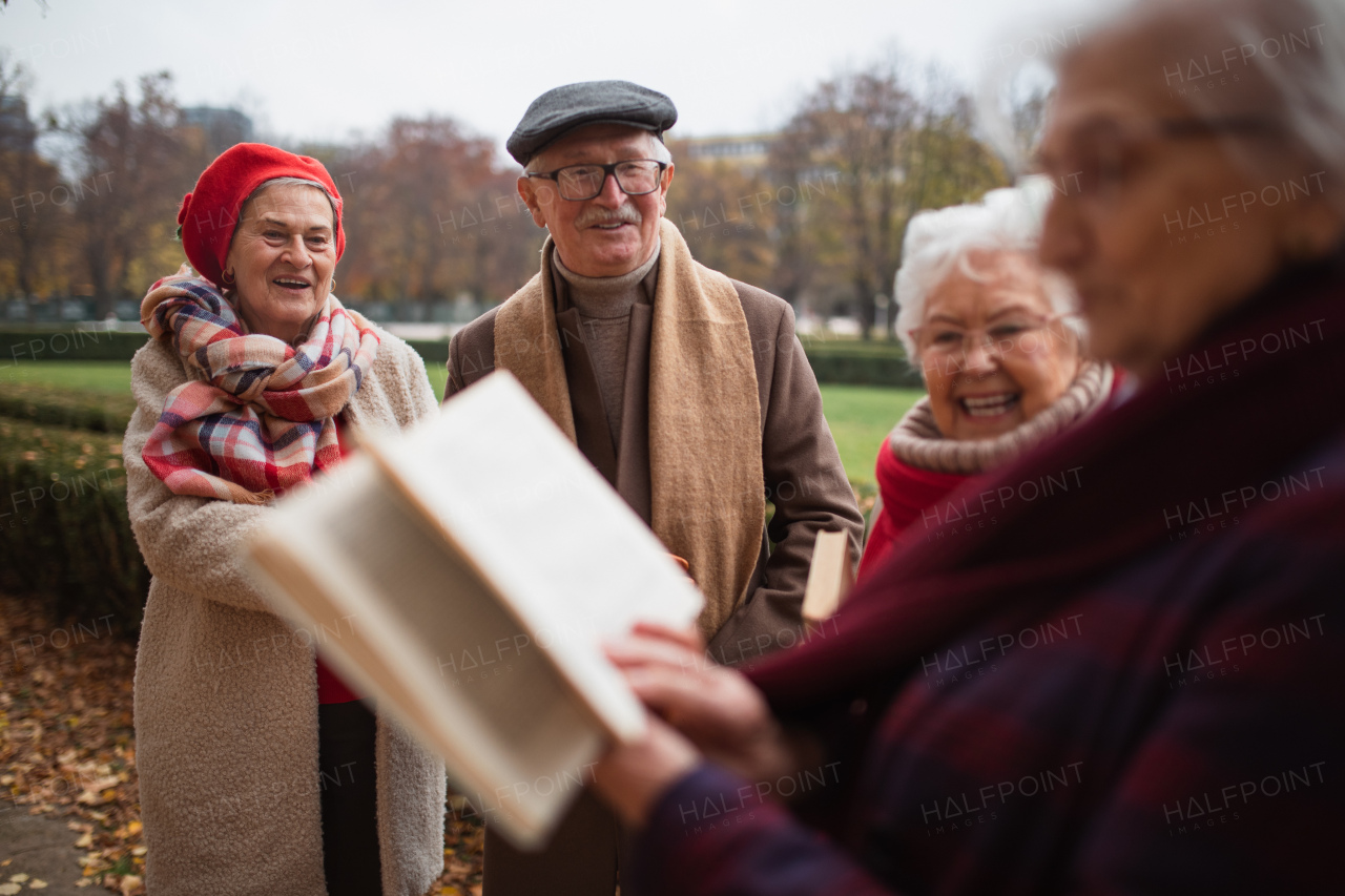 A group of happy senior friends with book on walk outdoors in park in autumn, reading and talking.