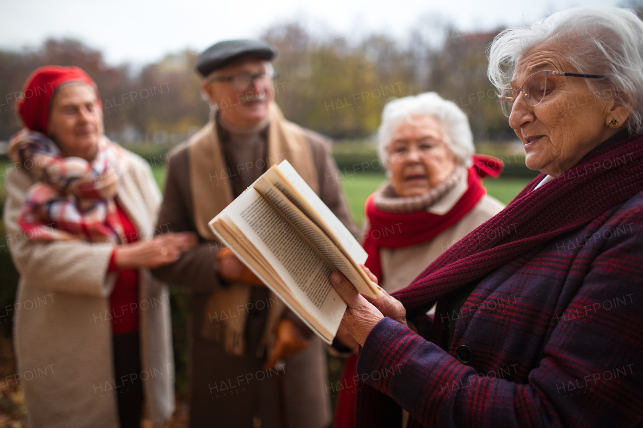 A group of happy senior friends with book on walk outdoors in park in autumn, reading and talking.