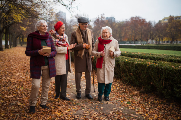 A group of happy senior friends with books on walk outdoors in park in autumn, talking and laughing