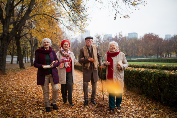 A group of happy senior friends with books on walk outdoors in park in autumn, talking and laughing