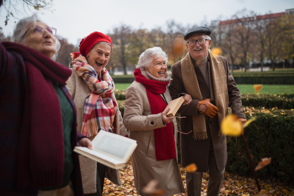 A group of happy senior friends with books on walk outdoors in park in autumn, talking and laughing