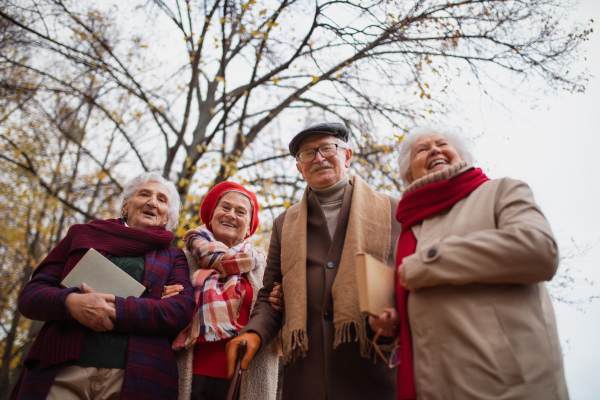 A low angle view of group of happy senior friends with books on walk outdoors in park in autumn