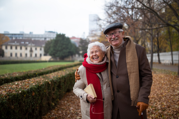 A happy senior couple on walk outdoors in park in autumn, embracing, laughing and looking at camera