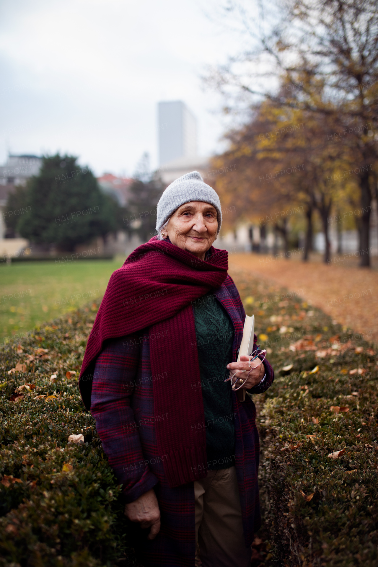 A happy senior woman with book on walk in town park in autumn, looking at camera.