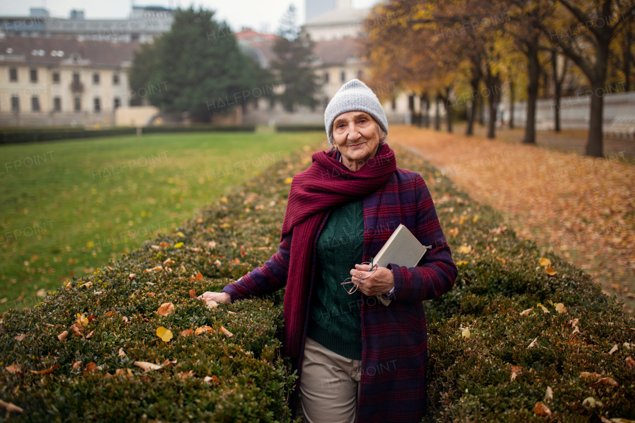 A happy senior woman with book on walk in town park in autumn, looking at camera.