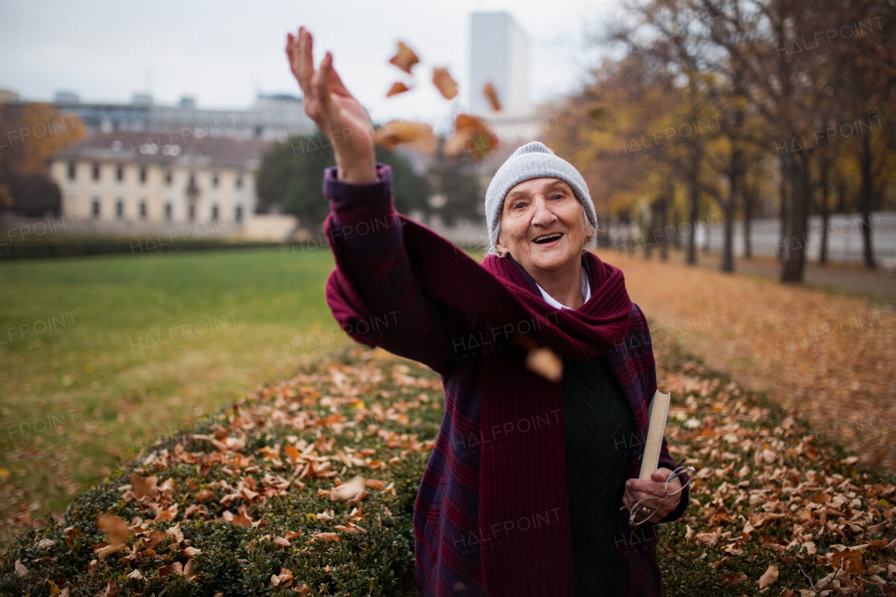 A happy senior woman on walk in town park in autumn, throwing leaves and looking at camera.