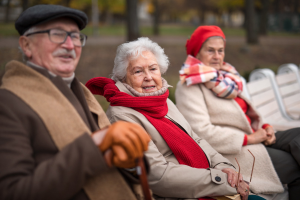 A group of happy senior friends sitting on bench in town park in autumn, looking at camera.