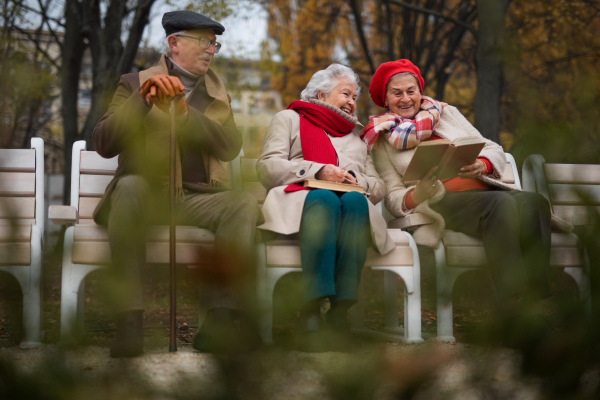 A group of happy senior friends sitting on bench in town park in autumn