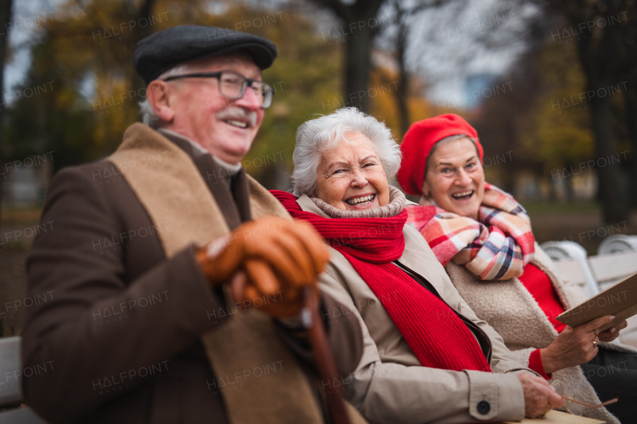 A group of happy senior friends sitting on bench in town park in autumn, looking at camera.