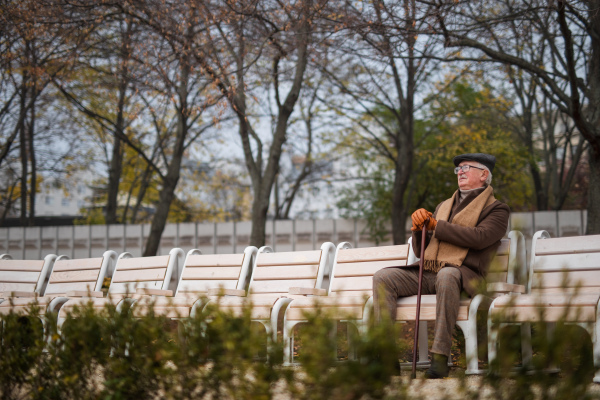 An old elegant man with walking stick sitting on bench in park on autumn day