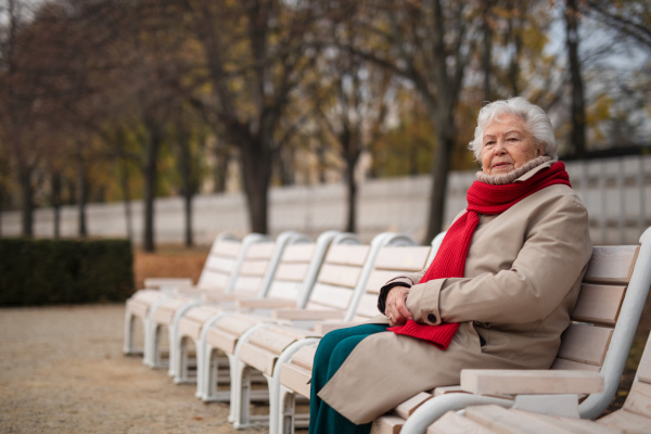 A senior woman sitting on bench in town park in autumn, looking at camera.