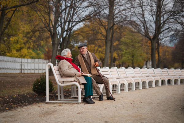 A happy senior friends sitting on bench and talking outdoors in park on autumn day.