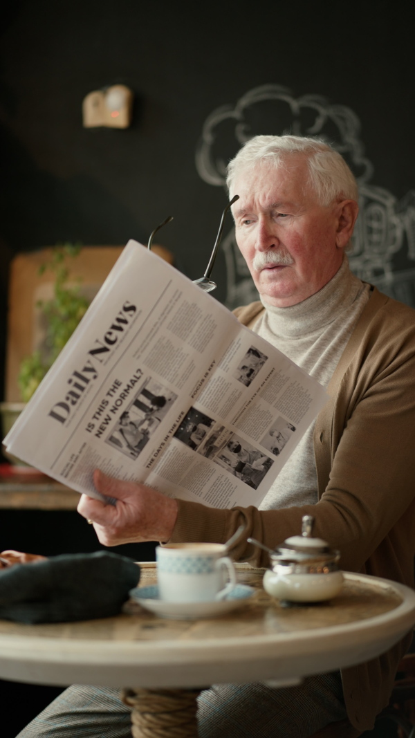 Elegant senior man with silver hair reading newspaper in a cafe, vertical footage.