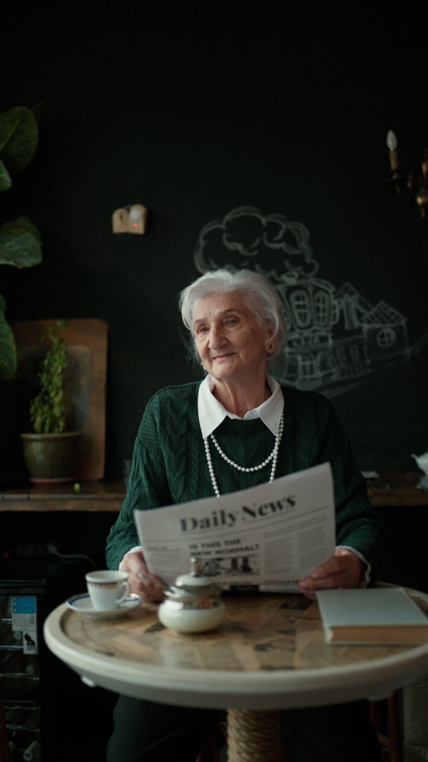 Elegant senior woman with silver hair and white pearls reading newspaper in cafe. Vertical footage.