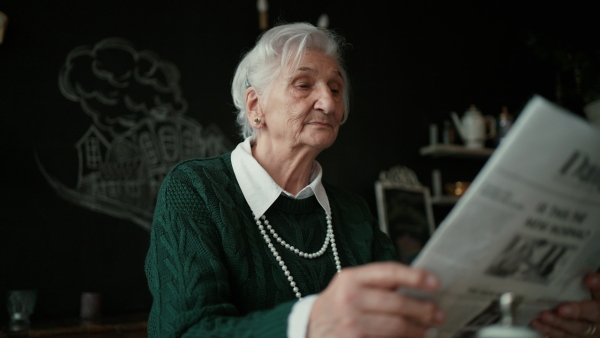 Elegant senior woman with silver hair and white pearls reading newspaper in cafe.