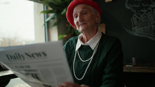 Elegant senior woman with silver hair and white pearls reading newspaper in cafe.