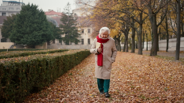 A happy senior woman with book on walk in town park in autumn.