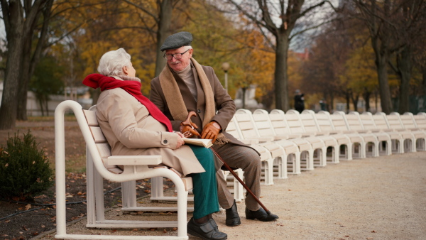 A happy senior friends sitting on bench and talking outdoors in park on autumn day.