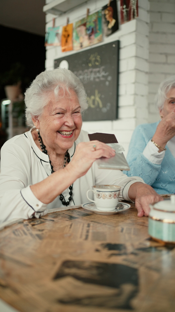 Happy senior friends sitting indoors in a cafe drinking coffee or tea, eating chocolate, laughing and having nice time together. Vertical footage.