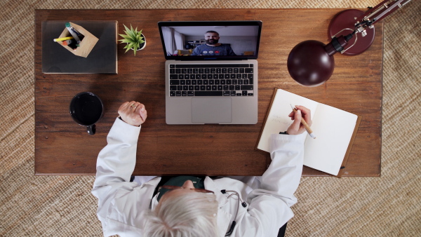 Top view of senior doctor having an online consultation on laptop, looking at camera. Lockdown concept.