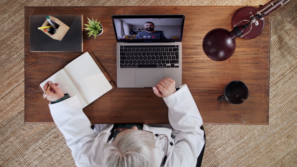 Top view of senior doctor having an online consultation on laptop, looking at camera. Lockdown concept.