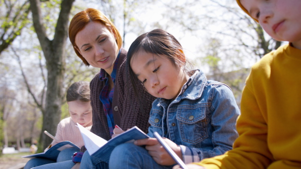 A teacher with small children sitting outdoors in city park, learning group education and coronavirus concept.