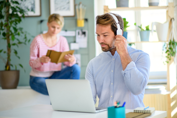 Mature businessman with headphones and laptop indoors in home office, working. Wife in the background.