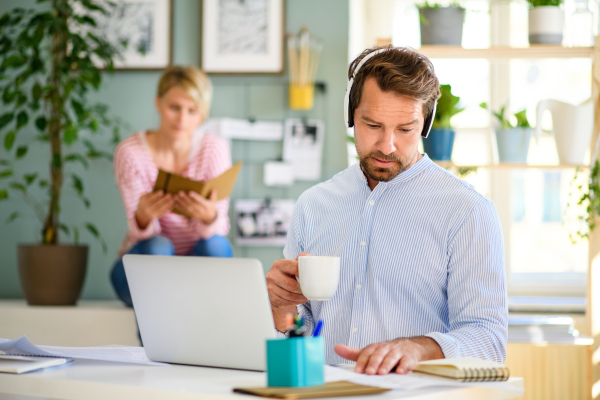 Front view of mature businessman with headphones and laptop indoors in home office, working.