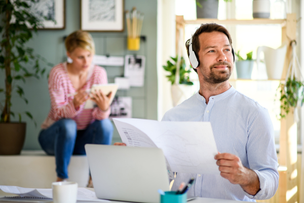 Mature architect with headphones, laptop and blueprints indoors in home office, working.