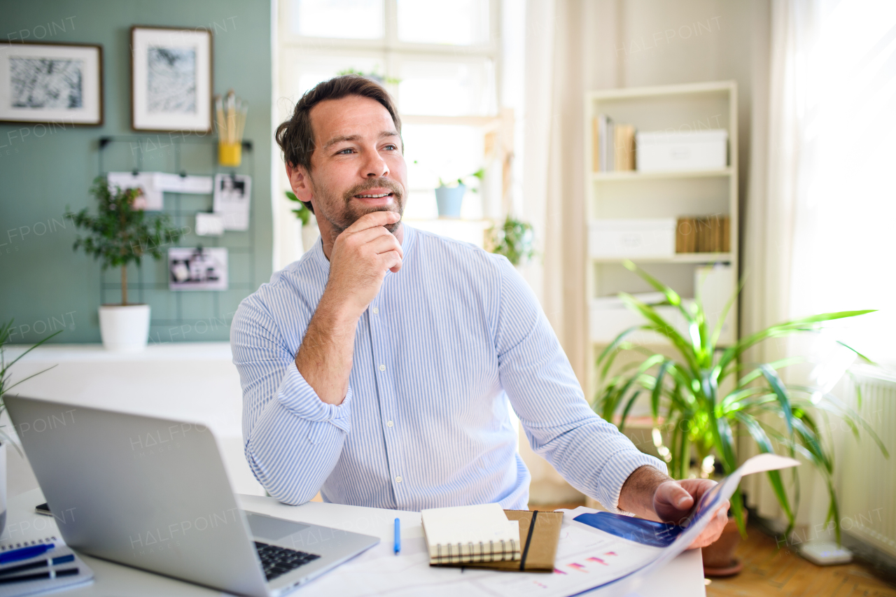 Front view of mature businessman with laptop indoors in home office, working.